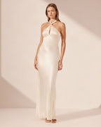 A woman modeling a floor-length satin halter gown in a warm ivory or cream tone against a soft beige studio backdrop with diagonal shadows.